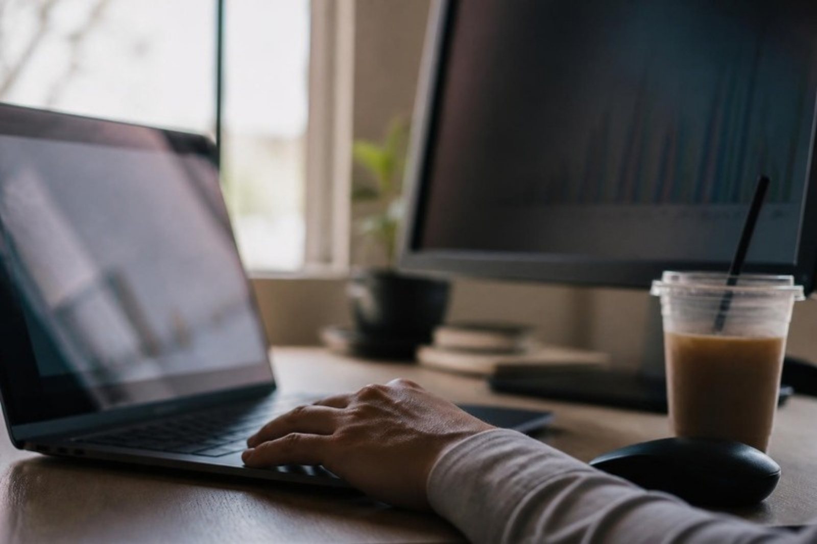 A hand resting on a laptop keyboard without typing, beside a second screen turned away showing an out-of-focus chart.